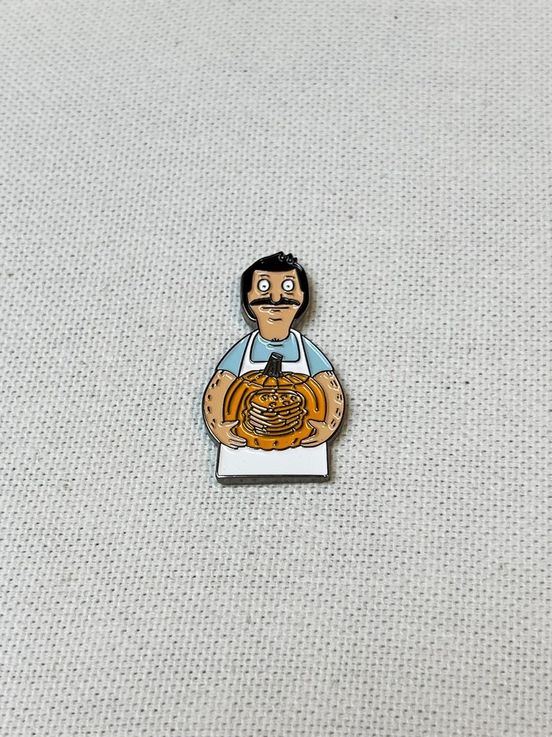 K&ouml;nnte beinhalten: Eine Emaille-Anstecknadel mit einem Cartoon-Mann mit Schnurrbart, der einen K&uuml;rbis h&auml;lt. Der K&uuml;rbis ist orange und hat ein geschnitztes Gesicht. Der Mann tr&auml;gt ein blaues Hemd und eine wei&szlig;e Sch&uuml;rze.