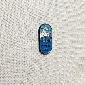 Op de afbeelding: Een blauwe en witte emaille pin in de vorm van een pil met een golf en een zeedier erin. De tekst "VITAMIN SEA" staat op de pin gedrukt.