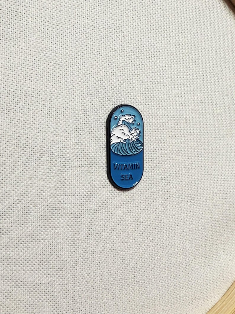 Op de afbeelding: Een blauwe en witte emaille pin in de vorm van een pil met een golf en een zeedier erin. De tekst "VITAMIN SEA" staat op de pin gedrukt.