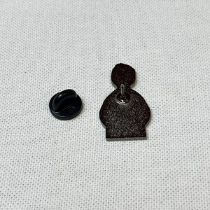 K&ouml;nnte beinhalten: Eine schwarze und silberne Emaille-Anstecknadel mit einer Cartoon-Figur mit schwarzem Umriss. Die Nadel hat einen Sicherheitsverschluss auf der R&uuml;ckseite.
