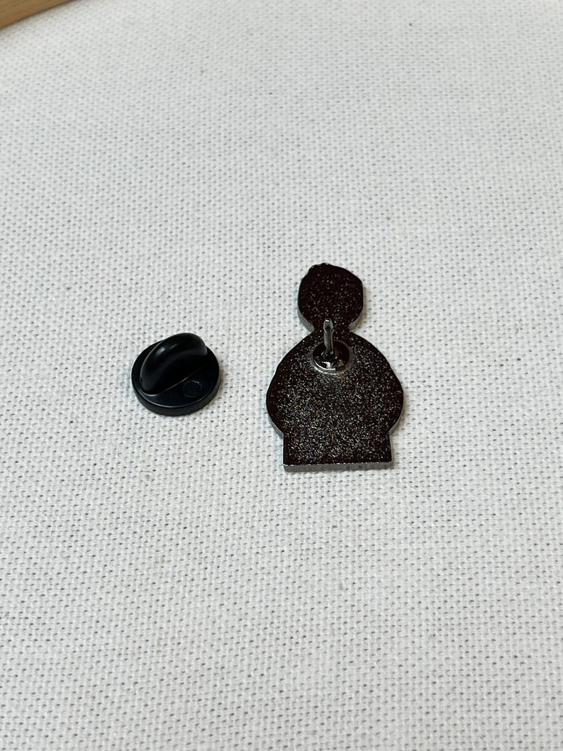 K&ouml;nnte beinhalten: Eine schwarze und silberne Emaille-Anstecknadel mit einer Cartoon-Figur mit schwarzem Umriss. Die Nadel hat einen Sicherheitsverschluss auf der R&uuml;ckseite.