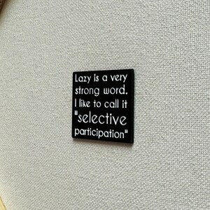 May include: Black square pin with white text that reads "Lazy is a very strong word. I like to call it "selective participation""