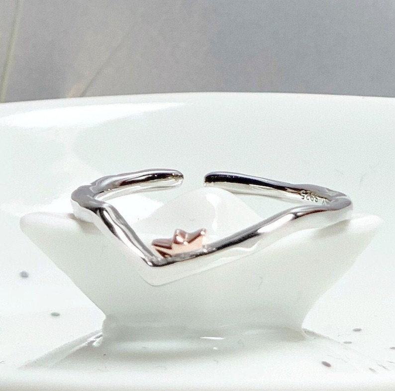 May include: A silver ring with a wavy design and a small rose gold crown on top.
