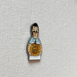 K&ouml;nnte beinhalten: Eine Emaille-Anstecknadel mit einem Cartoon-Mann mit Schnurrbart, der einen K&uuml;rbis h&auml;lt, der mit einem Stapel Pfannkuchen geschnitzt ist. Der Mann tr&auml;gt ein blaues Hemd und eine wei&szlig;e Sch&uuml;rze.