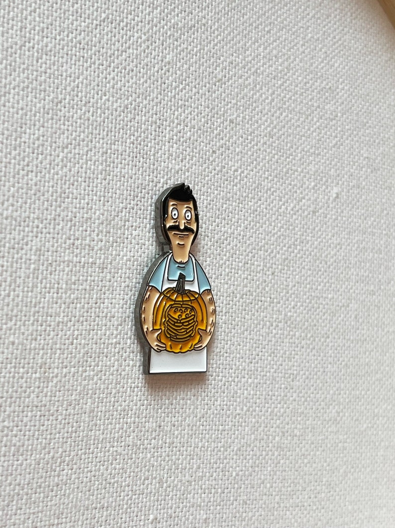K&ouml;nnte beinhalten: Eine Emaille-Anstecknadel mit einem Cartoon-Mann mit Schnurrbart, der einen K&uuml;rbis h&auml;lt, der mit einem Stapel Pfannkuchen geschnitzt ist. Der Mann tr&auml;gt ein blaues Hemd und eine wei&szlig;e Sch&uuml;rze.