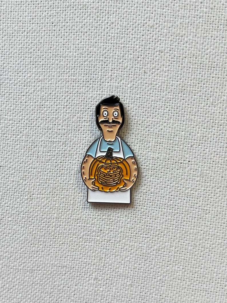 K&ouml;nnte beinhalten: Eine Emaille-Anstecknadel mit einem Cartoon-Mann, der einen K&uuml;rbis mit einem Stapel Pfannkuchen h&auml;lt. Der Mann tr&auml;gt ein blaues Hemd und eine wei&szlig;e Sch&uuml;rze. Der K&uuml;rbis ist orange und die Pfannkuchen sind braun.