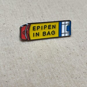 K&ouml;nnte beinhalten: Gelbe und rote Emaille-Anstecknadel in Form eines Epipens mit dem Text "EPIPEN IN BAG" darauf.