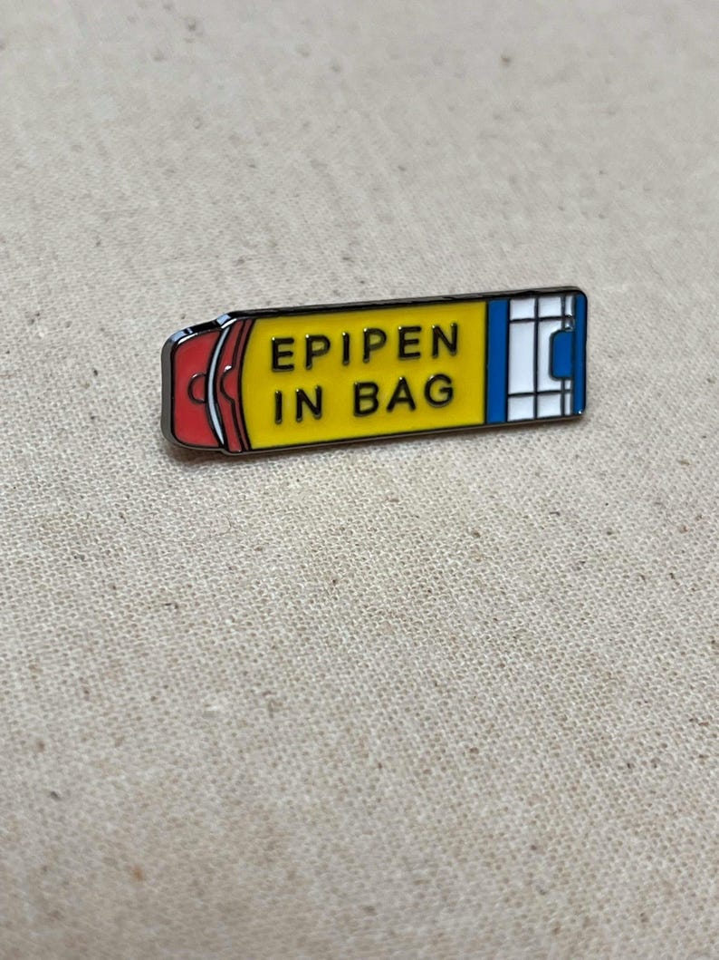 K&ouml;nnte beinhalten: Gelbe und rote Emaille-Anstecknadel in Form eines Epipens mit dem Text "EPIPEN IN BAG" darauf.