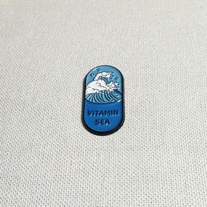 Op de afbeelding: Een blauwe en witte emaille pin in de vorm van een pil met een golfontwerp en de tekst "VITAMIN SEA".
