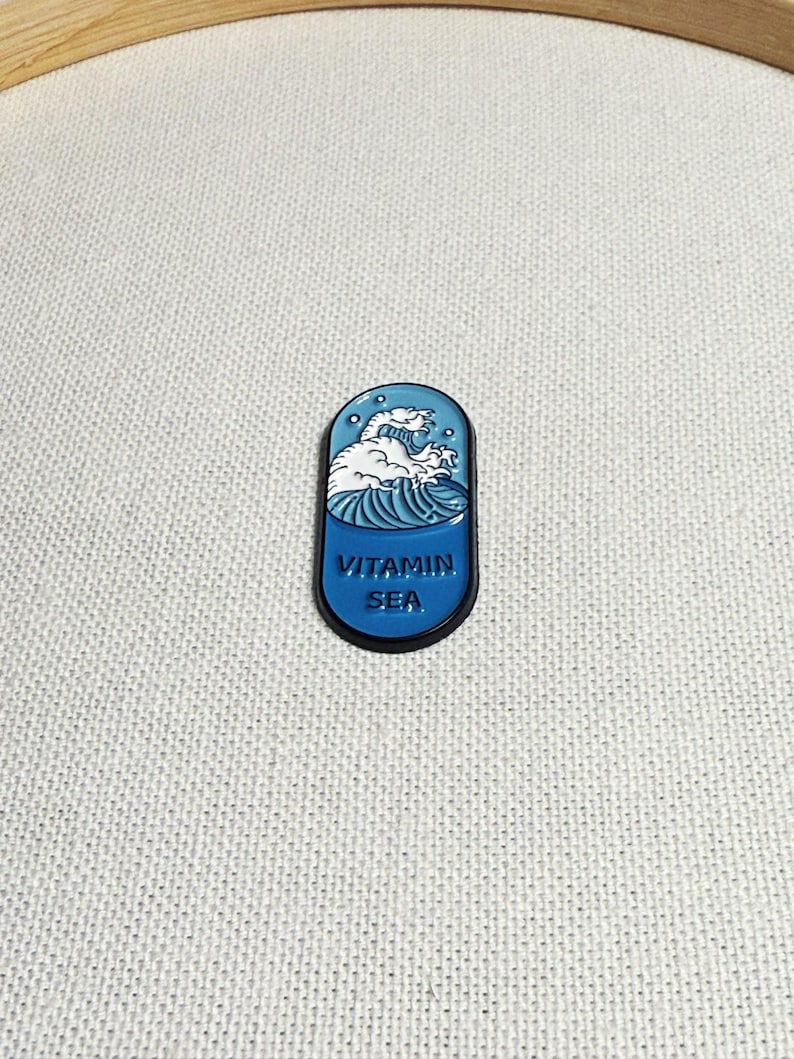 Op de afbeelding: Een blauwe en witte emaille pin in de vorm van een pil met een golfontwerp en de tekst "VITAMIN SEA".