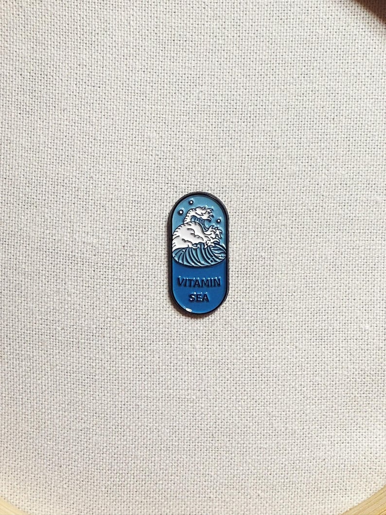 Op de afbeelding: Een blauwe en witte emaille pin in de vorm van een pil met een golf en de tekst "Vitamin Sea" erop.