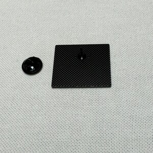 May include: A black enamel pin with a textured surface. The pin is on a white fabric background.