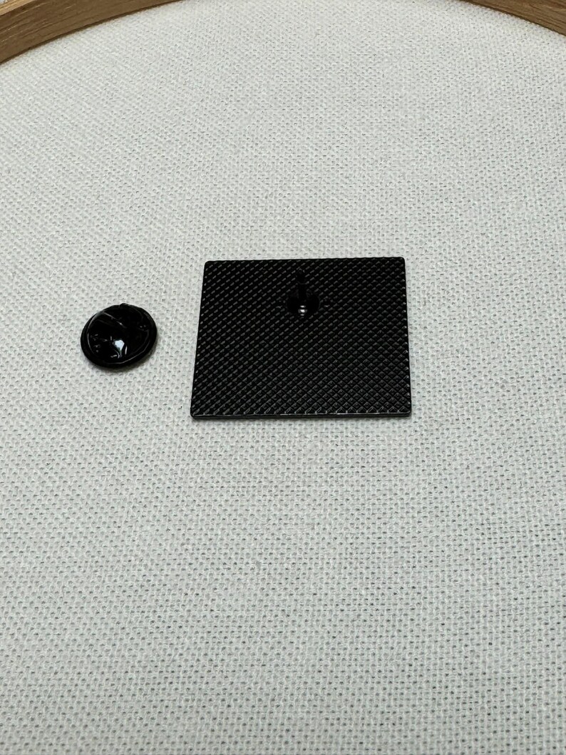 May include: A black enamel pin with a textured surface. The pin is on a white fabric background.