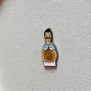 K&ouml;nnte beinhalten: Eine Metallstift mit einer Zeichentrickfigur, die einen K&uuml;rbis h&auml;lt. Die Figur tr&auml;gt ein blaues Hemd und eine wei&szlig;e Sch&uuml;rze. Der K&uuml;rbis ist orange und hat ein geschnitztes Gesicht.