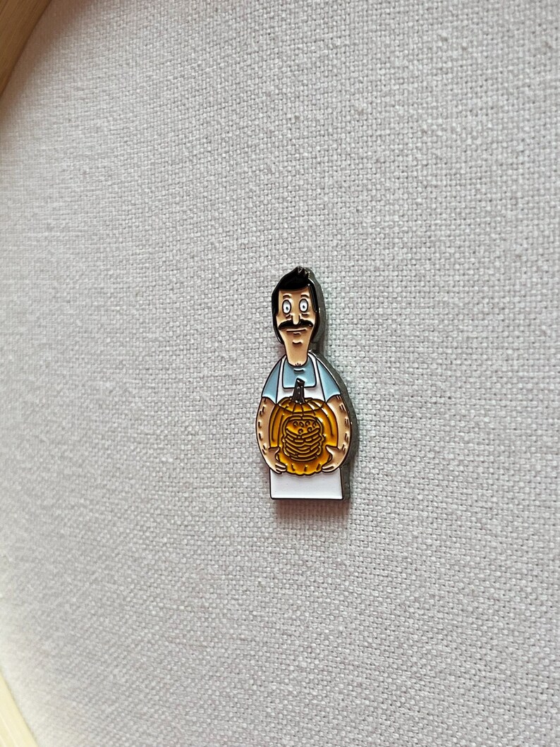 K&ouml;nnte beinhalten: Eine Metallstift mit einer Zeichentrickfigur, die einen K&uuml;rbis h&auml;lt. Die Figur tr&auml;gt ein blaues Hemd und eine wei&szlig;e Sch&uuml;rze. Der K&uuml;rbis ist orange und hat ein geschnitztes Gesicht.