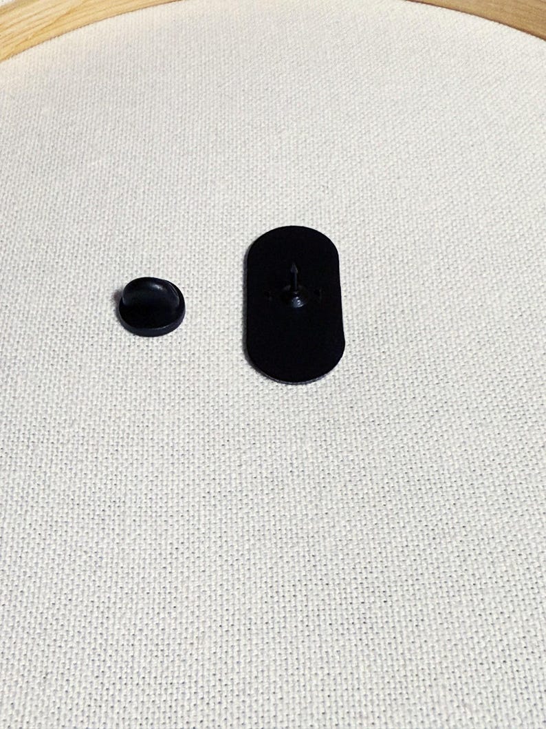 Op de afbeelding: Twee zwarte metalen pinruggen voor een speld of badge. De grotere pinrug is ovaalvormig en de kleinere is rond.