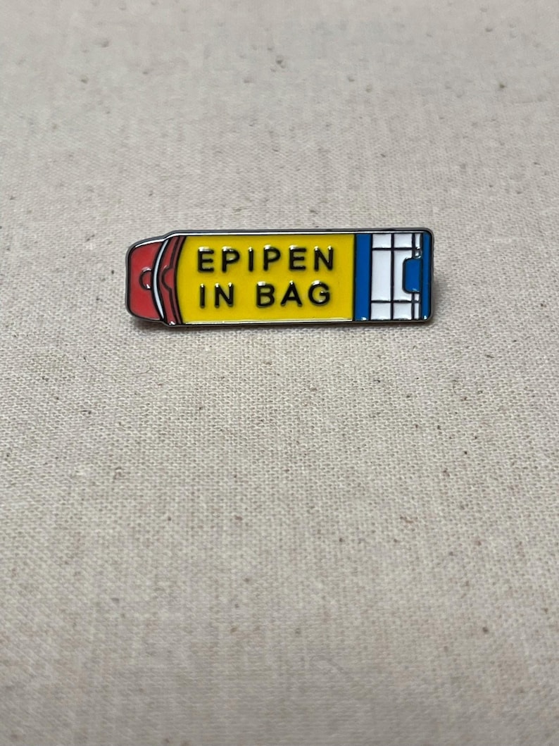 K&ouml;nnte beinhalten: Eine gelbe und blaue Emaille-Anstecknadel in Form eines Epipens mit dem Text "EPIPEN IN BAG" in schwarzen Buchstaben.