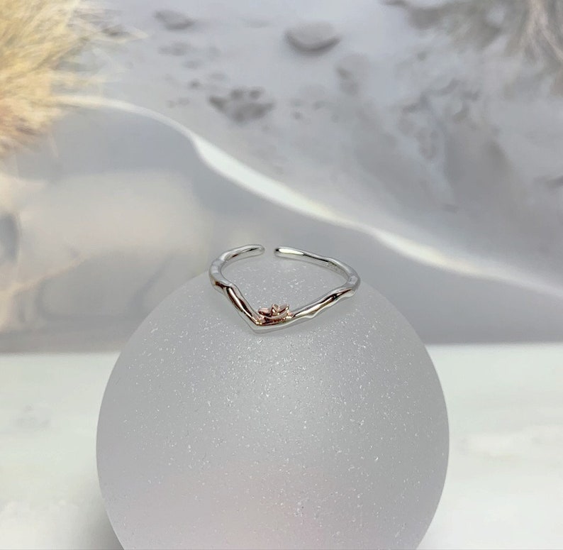 May include: A silver ring with a delicate, uneven, V-shaped design. The ring has a small, rose gold flower accent in the center.