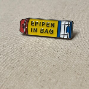 K&ouml;nnte beinhalten: Eine gelbe und blaue Emaille-Anstecknadel in Form eines Epipens mit dem Text "EPIPEN IN BAG" darauf.