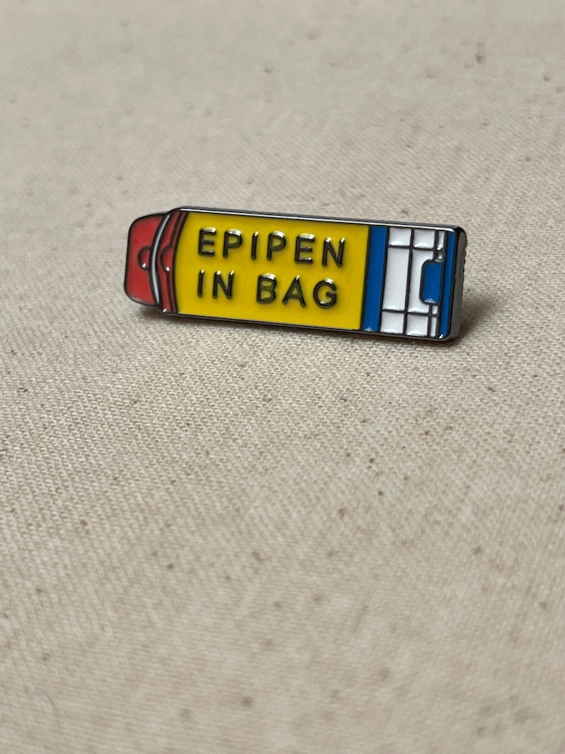 K&ouml;nnte beinhalten: Eine gelbe und blaue Emaille-Anstecknadel in Form eines Epipens mit dem Text "EPIPEN IN BAG" darauf.