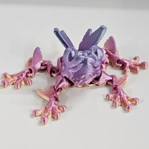 May include: A 3D printed frog figurine with a purple and pink gradient color scheme. The frog has large, outstretched legs and a detailed, textured surface.
