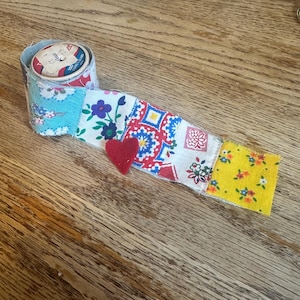 Può includere: Un nastro patchwork colorato con un cuore di feltro rosso. Il nastro è fatto di diversi tessuti con motivi floreali e geometrici.