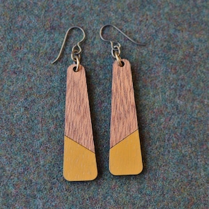 May include: A pair of long, wooden earrings with a geometric design. The earrings are painted in two shades of brown and yellow.