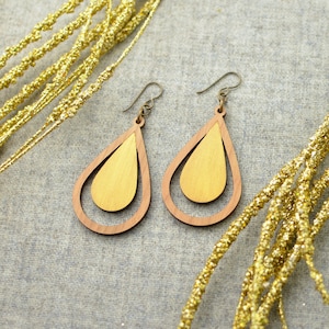 May include: A pair of wooden teardrop earrings with a gold finish. The earrings are made of two layers of wood, with the top layer being a lighter shade of wood than the bottom layer.