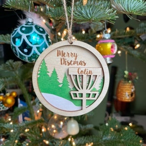 May include: A wooden Christmas ornament shaped like a disc golf basket with a green forest scene and the text "Merry Discmass Colin".