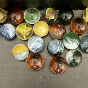 Puede incluir: Una colección de canicas de vidrio de colores en varios tamaños y diseños. Las canicas presentan una gama de colores, incluyendo verde, amarillo, rojo, azul y blanco, con patrones como remolinos y colores sólidos. Las canicas están dispuestas sobre una superficie oscura.