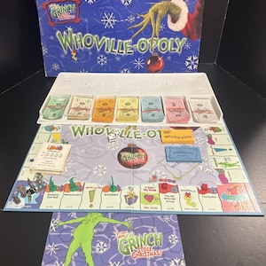 May include: A Whoville-Opoly board game set featuring a blue box with a green Grinch hand, a game board with snowflake patterns, colorful paper money in a white tray, metal game tokens, dice, red house markers, and a rule booklet.