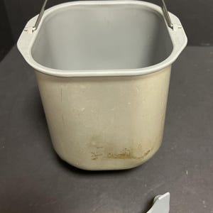 May include: A beige bread machine bucket with a silver handle and a detached grey paddle. The bucket has a square shape with rounded corners and a grey interior. The paddle is lying on a dark surface.