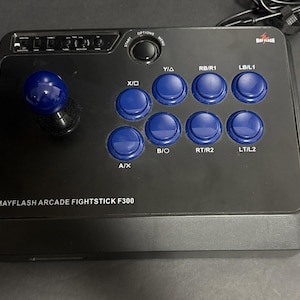 May include: A black Mayflash Arcade Fightstick F300 controller featuring a blue joystick and eight blue action buttons labeled with console-style inputs. The top panel includes a start button, options button, and a row of small toggle switches.