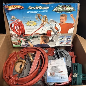 May include: A Hot Wheels AcceleDrome track set in a cardboard box. The packaging features the text AcceleDrome and Acceleracers with images of a motorized booster, rotating loops, and a child playing. Inside are red plastic track pieces.