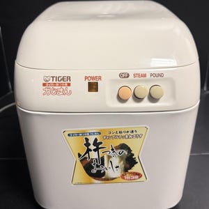 Może przedstawiać: Białe urządzenie do robienia ciasta ryżowego Tiger z japońskim tekstem i przyciskami sterowania. Urządzenie posiada przycisk zasilania oraz przyciski do funkcji wyłączania, gotowania na parze i ubijania. Naklejka z przodu zawiera japoński tekst i ilustracje.