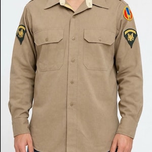 Op de afbeelding: Een beige overhemd met lange mouwen, knoopsluiting, twee borstzakken, een lichtcrèmekleurige binnenkraag en patches in militaire stijl op beide schouders. Het overhemd wordt gedragen met een blauwe spijkerbroek voor een gestructureerde uitstraling.