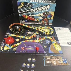 May include: Zathura board game with a space theme. The game includes a game board, game pieces, and a box with the text "Zathura Adventure is Waiting". The game board features a track with a black ball, a red sphere, and a purple control panel.