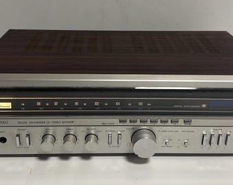 May include: A Sansui digital synthesiser DC stereo receiver with a silver metal front panel, multiple control knobs, and push-button switches. The top casing features a dark wood grain finish. The display shows frequency scales and the Sansui logo.