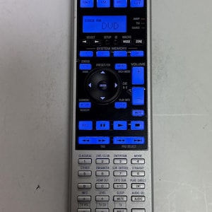 Pode incluir: Controle remoto prateado Yamaha RAV389 com botões retroiluminados azuis. Possui botões para DVD, TV e controle de volume. A palavra "DVD" é exibida na tela.