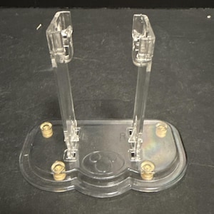 Könnte beinhalten: Ein transparenter Acryl-Präsentationsständer mit zwei vertikalen Stützen auf einer ovalen Basis. Die Basis verfügt über vier kleine goldfarbene Metallfüße und ein mittig eingeprägtes kreisförmiges Symbol. Die Buchstaben L und R sind eingeprägt.