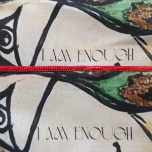 May include: Close-up of a fabric with a repeating pattern of stylized eyes and abstract shapes in black, green, and beige. The text "I AM ENOUGH" is printed in black. A red seam runs across the image.