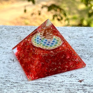 Può includere: Piramide di cristallo rosso con una bobina di filo di rame e un disegno di fiore della vita sulla cima. La piramide è fatta di cristalli rossi e ha una finitura trasparente e lucida.