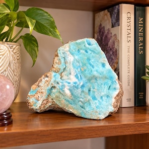 Peut inclure: Un morceau brut et irrégulier de minéral hémimorphite bleu vif avec une surface cristalline texturée. Le spécimen présente une coloration bleu turquoise éclatante contrastée par des taches de matrice terreuse beige et brune sur ses bords.