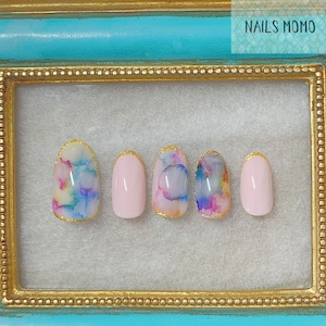 May include: Five oval-shaped false nails with a pink base and gold trim. Two nails feature a watercolour design with blue, pink, and yellow colours. The nails are displayed in a gold frame with a turquoise background. The text "NAILS MOMO" is visible in the top right corner of the image.