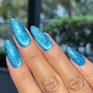 Puede incluir: Primer plano de una mano con uñas largas en forma de almendra. Las uñas están pintadas de un azul vibrante y brillante, con un acabado brillante que captura la luz. El tono de piel es cálido y medio, contrastando con el color brillante de las uñas.