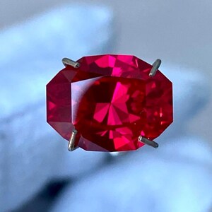 May include: A deep red gemstone with a faceted cut, held in place by four prongs. The gemstone is oval shaped and has a high polish.