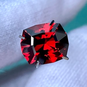 May include: A deep red gemstone with a faceted cut. The gemstone is held in place by four prongs.