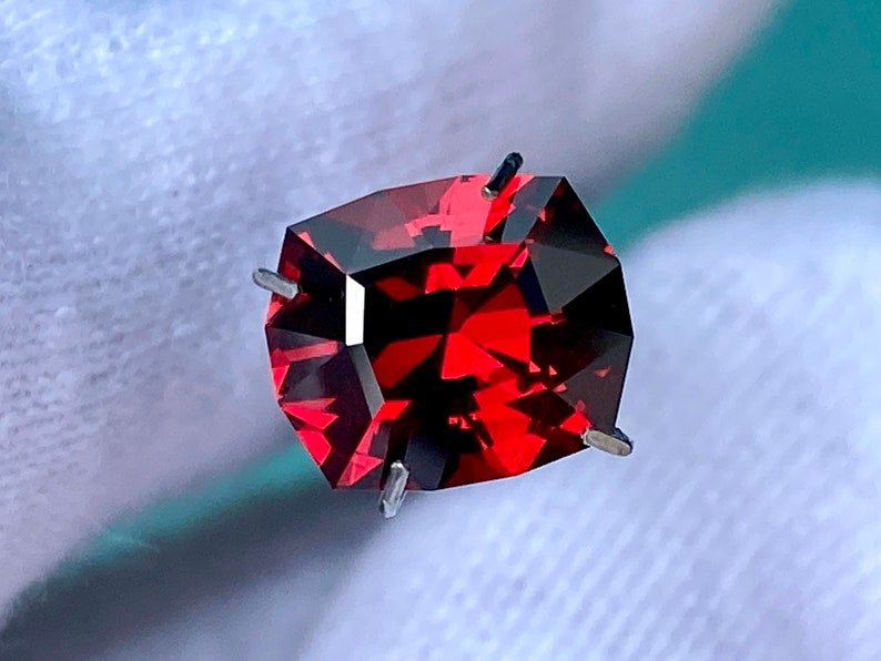 May include: A deep red gemstone with a faceted cut. The gemstone is held in place by four prongs.