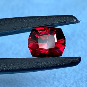 May include: A deep red gemstone with a faceted octagonal shape, held by tweezers against a blue background.