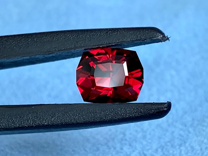 May include: A deep red gemstone with a faceted octagonal shape, held by tweezers against a blue background.